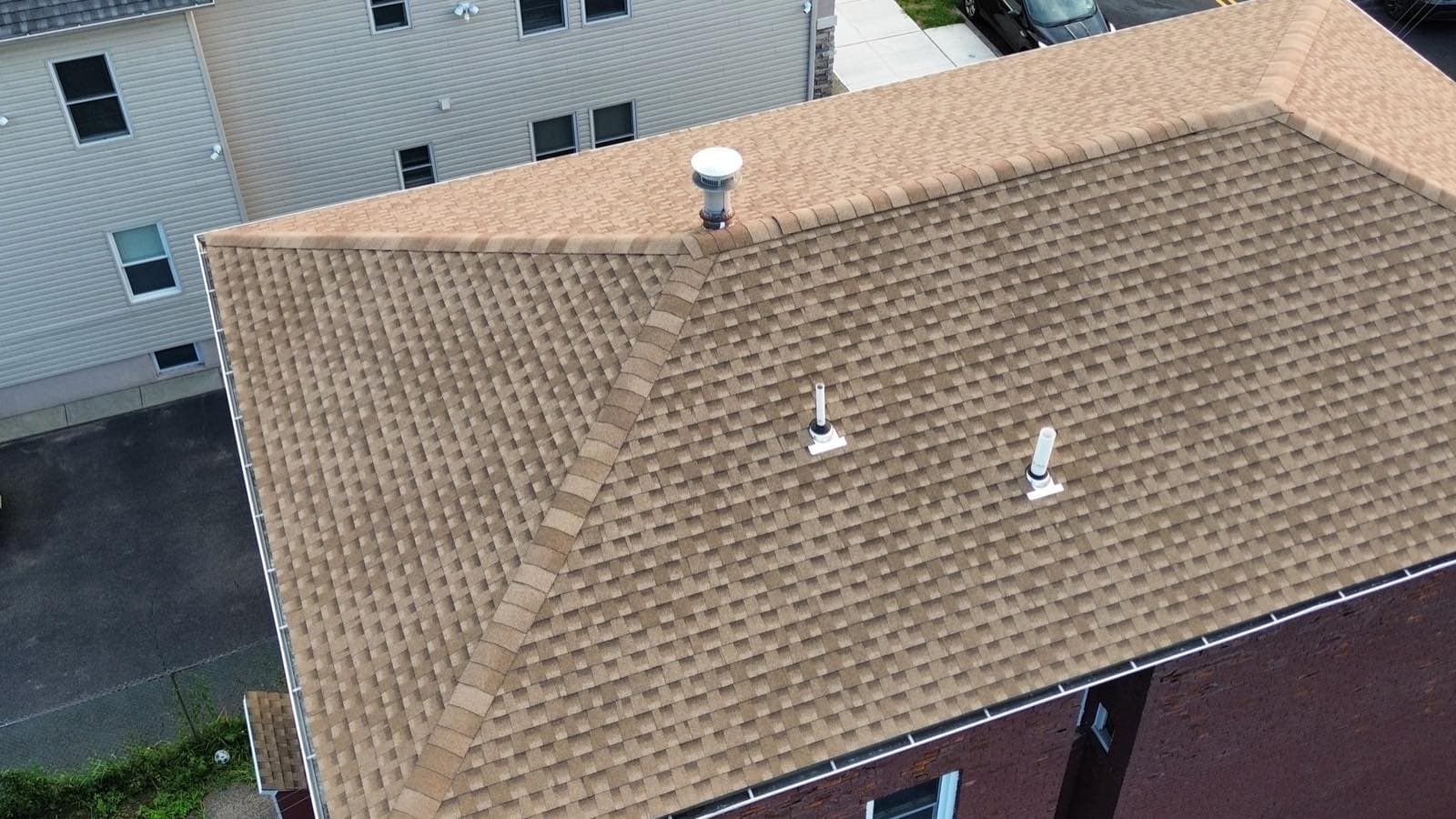 Close aerial of the completed Lodi, NJ hip roof showing capped hips, a ridge vent and two flashed pipe boots set into the shingle field