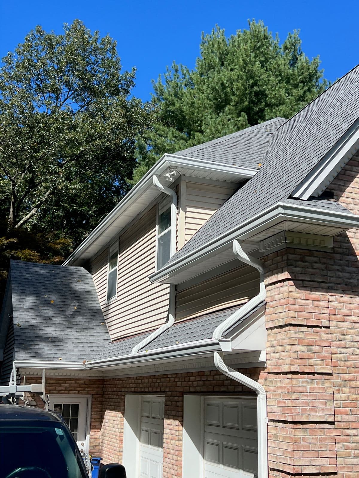 Gray architectural asphalt shingle roof with new white gutters and downspouts over the garage wing and dormers of a brick and vinyl-sided home in Franklin Lakes, NJ, with a pickup truck with a ladder rack in the foreground