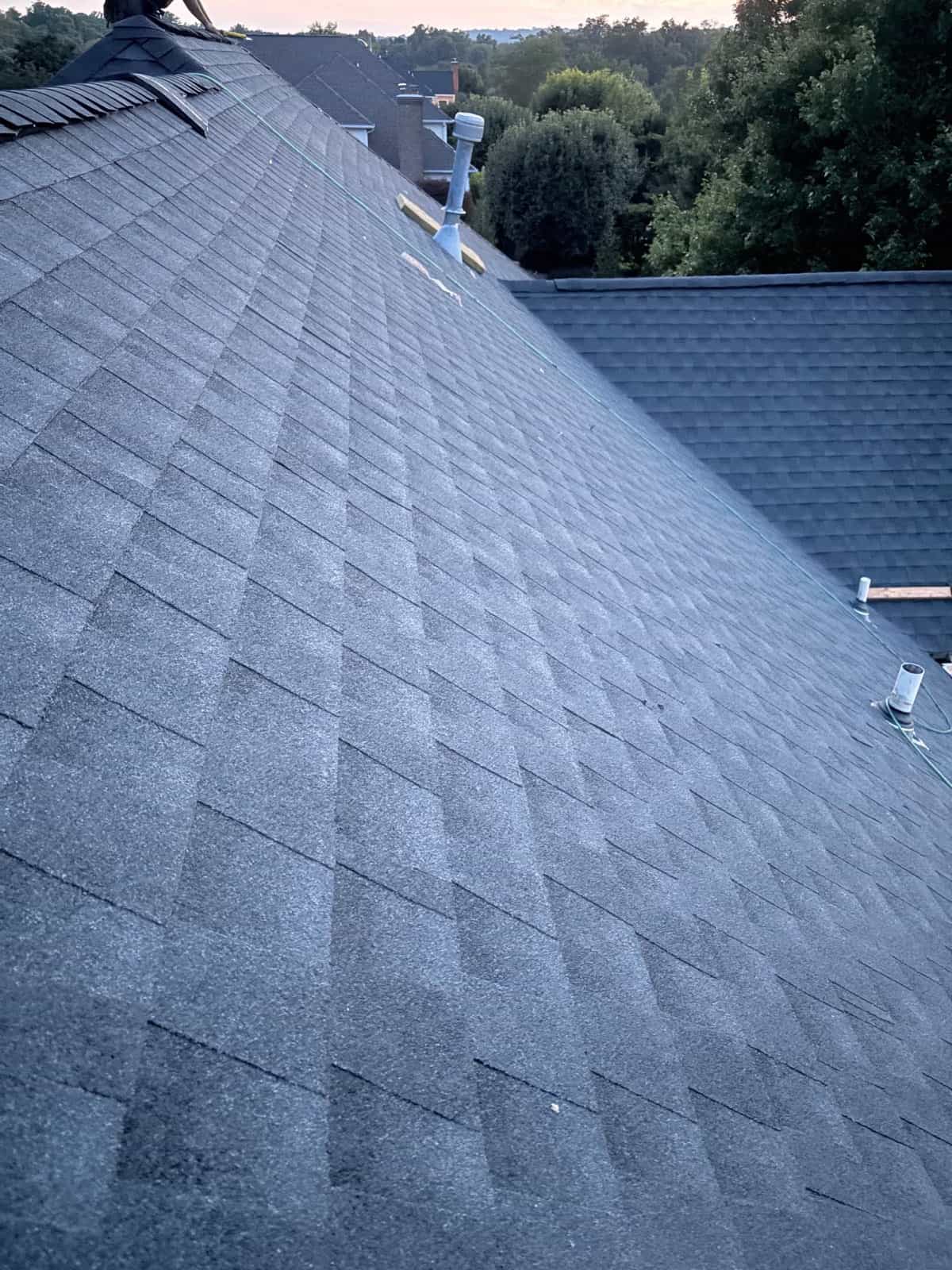 Close view of a dark gray asphalt-shingle roof slope running up to the ridge, with a pipe vent and a neighboring roof beyond, at dusk
