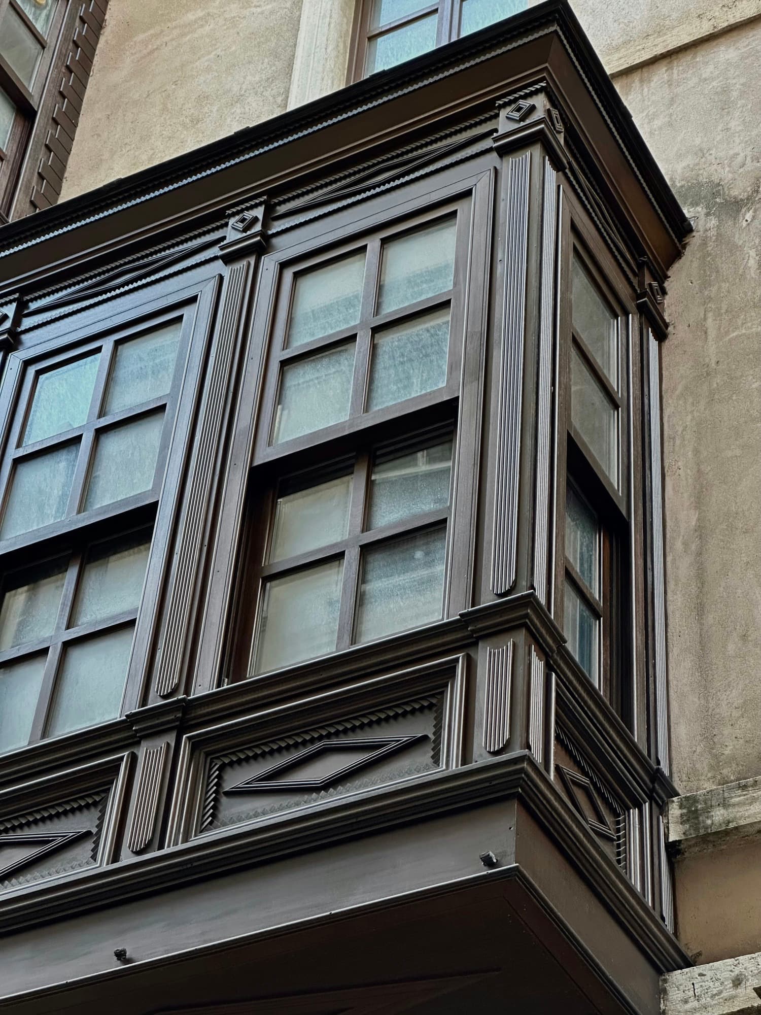 Dark-stained wooden bay window with divided-lite sashes projecting from a stucco building