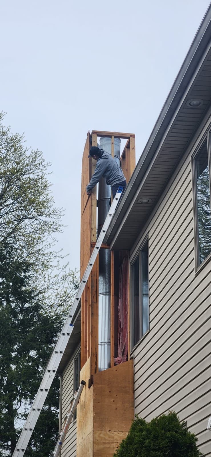 Carpenter rebuilding a framed chimney chase on a New Jersey home, new studs and sheathing going up around the existing metal flue where the chase passes the roof edge