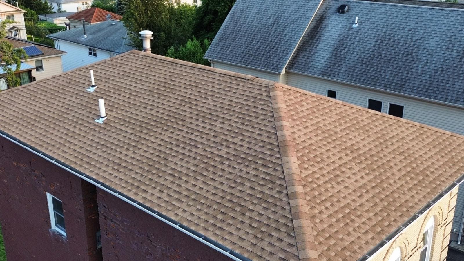 Completed tan architectural shingle hip roof on a brick two-family in Lodi, NJ, with new white seamless gutters run along the eave