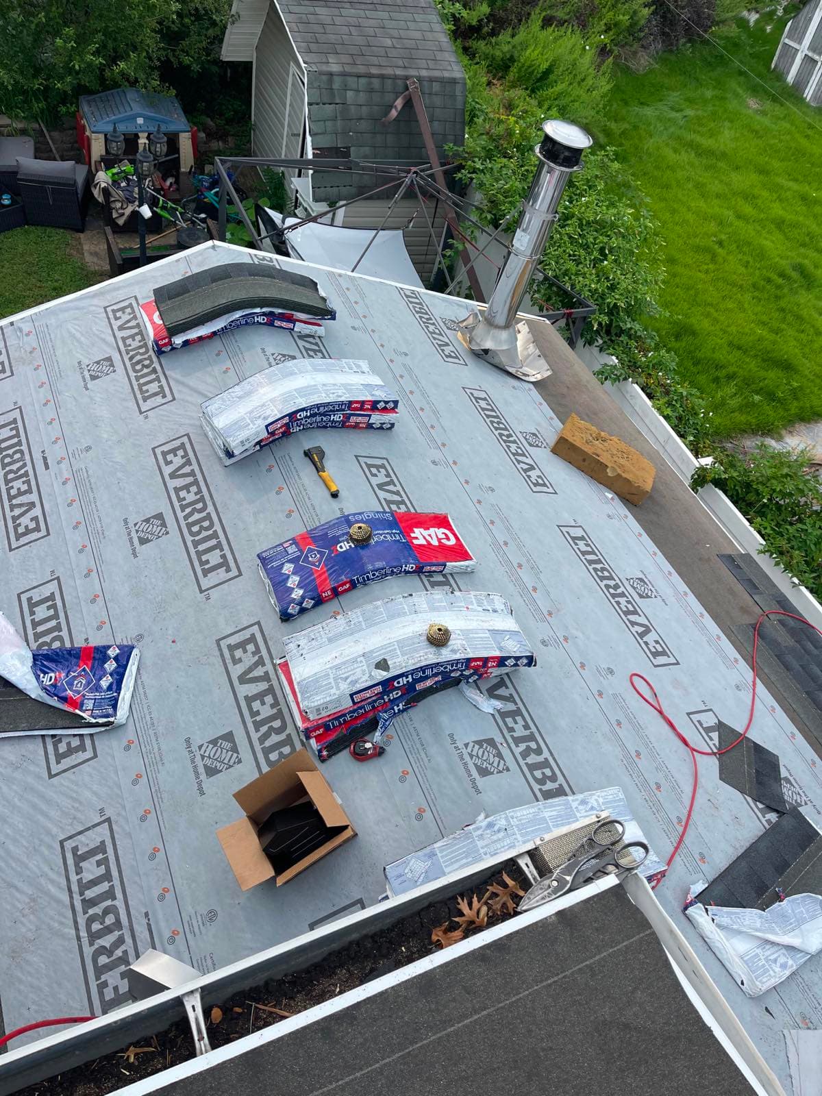 GAF Timberline HD shingle bundles staged in rows across synthetic underlayment on a Parlin, NJ roof, with a stainless flue pipe flashed in and coil nailers set out ready for the field