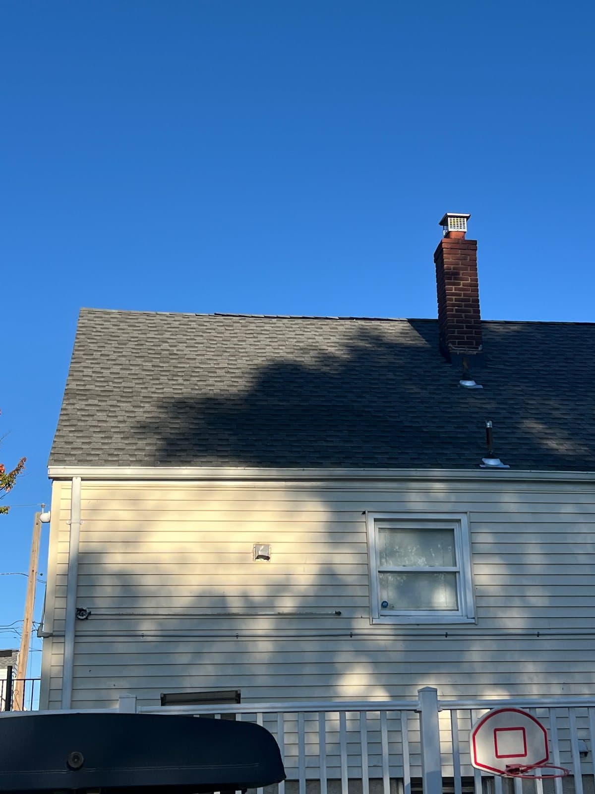 Back of a house in Passaic, NJ with a new asphalt shingle roof in dappled tree shade and a brick chimney with a metal cap rising from the slope