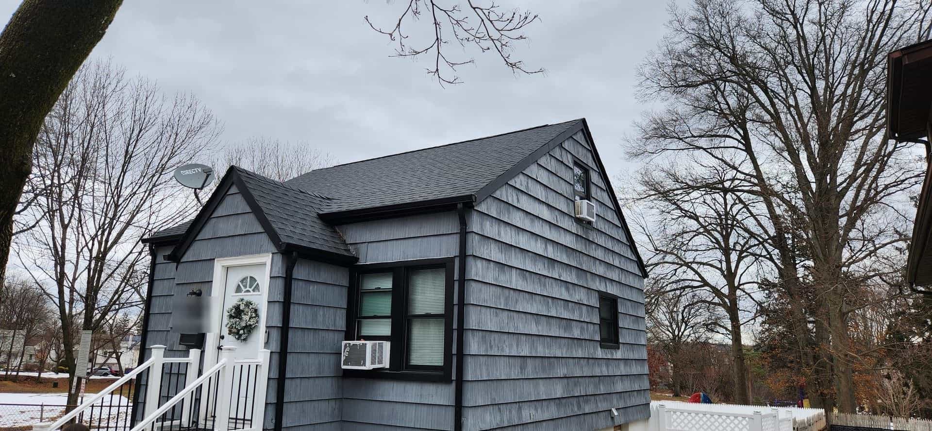 Gray-shingled Cape Cod house with a wreath on the front door, a dark roof and gutters, and bare winter trees