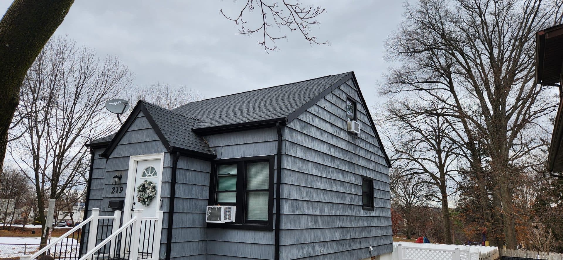 Cape cod in Teaneck, New Jersey after a full roof replacement in dark architectural asphalt shingle, with new black gutters, downspouts and fascia trim running the eaves and both gable rakes