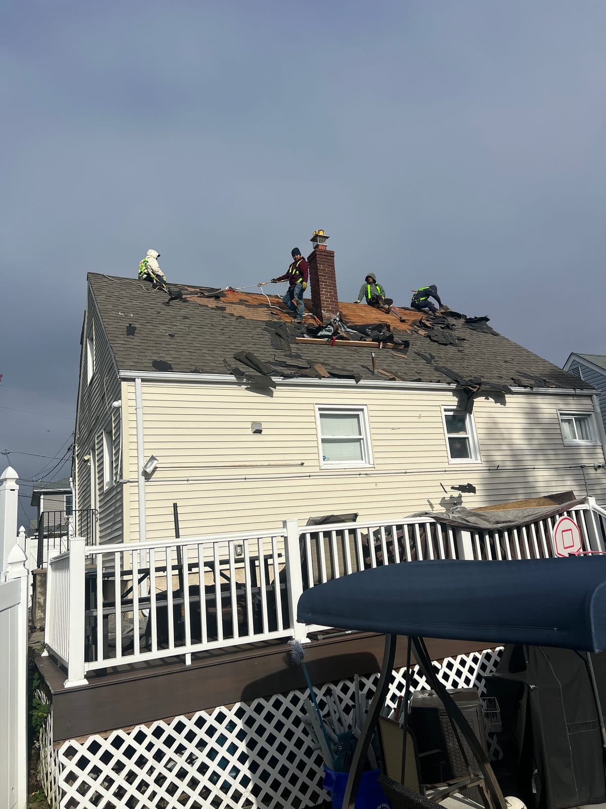 Roofers tied off with safety harnesses and lines on a Passaic, NJ roof, old shingles removed to the wood deck beside a brick chimney, seen from the back deck