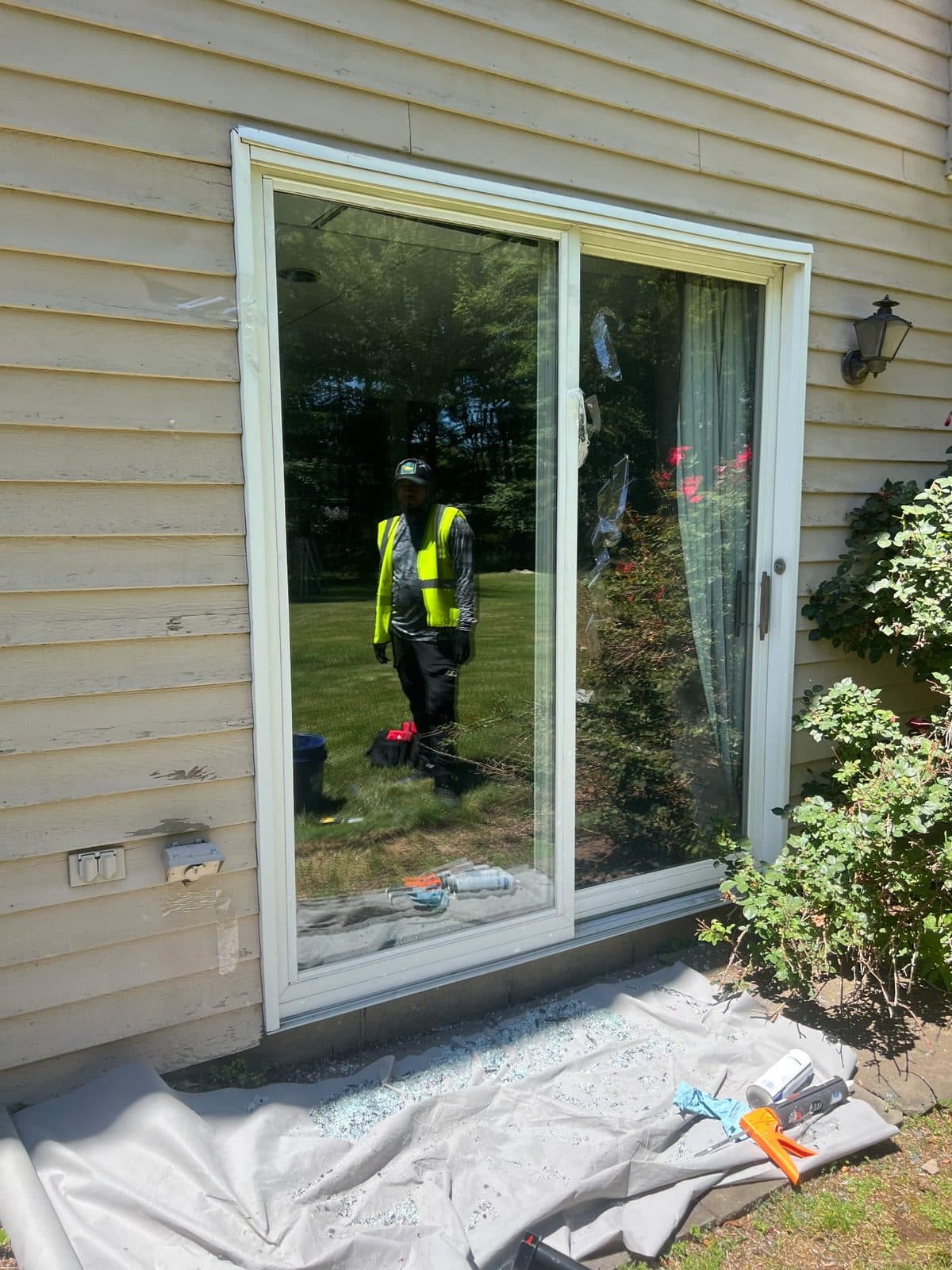 Precision technician replacing the fixed glass panel of a sliding patio door on a Clifton, NJ home — shattered tempered glass cleared onto a drop cloth below the new pane