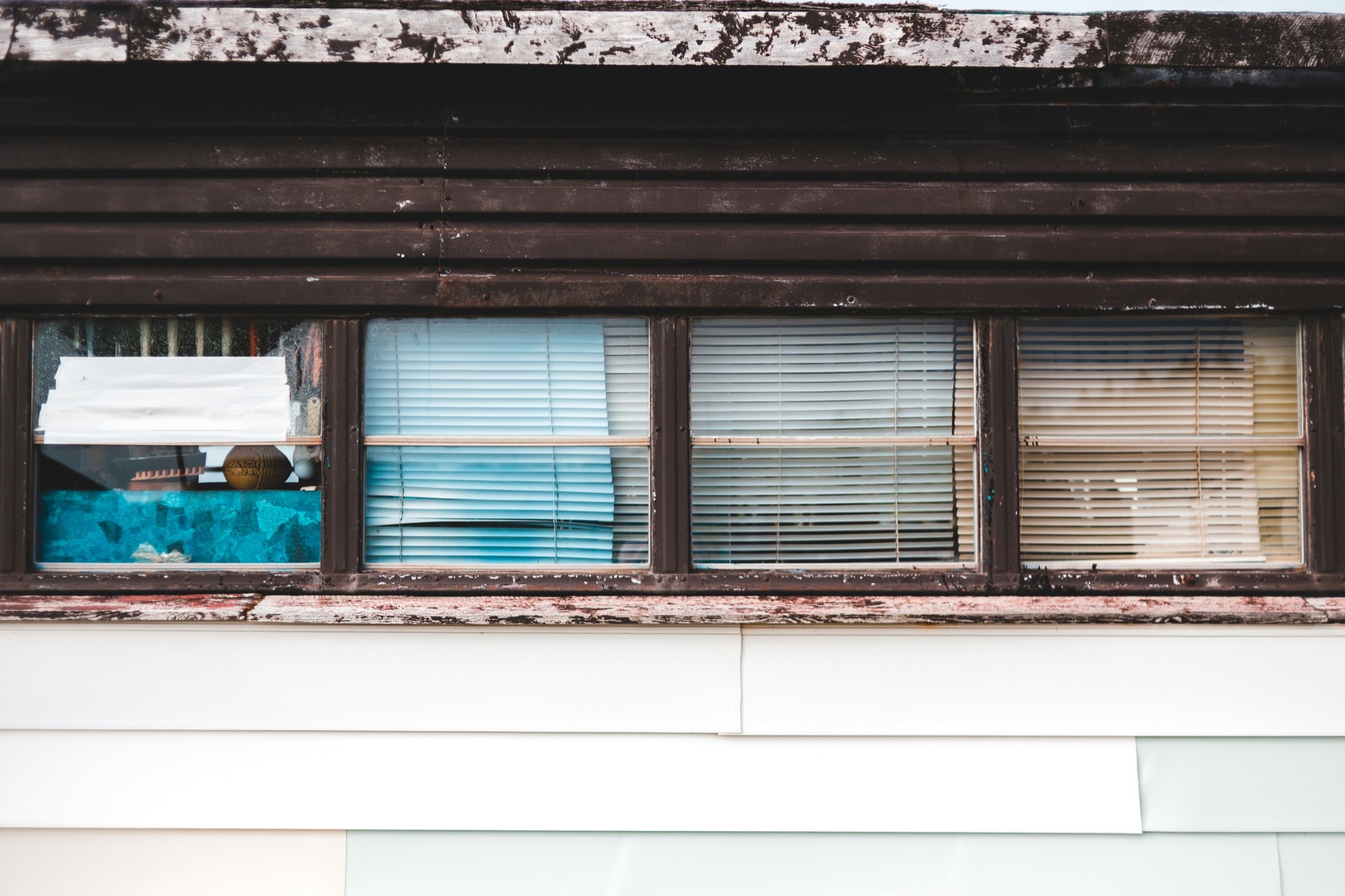 Row of weathered dark-framed windows with blinds behind the glass, above white siding